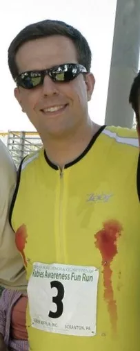 Smiling man in sunglasses and yellow running shirt with red stains on the chest, wearing race bib number 3