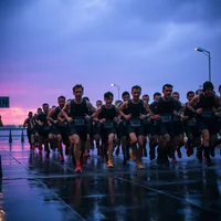 Large pack of soaked military runners in monochrome kits crossing a rainy waterfront promenade finish line at twilight, shot from ground level.