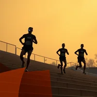 Three silhouetted runners ascending outdoor stadium stairs against a vivid orange-golden sky
