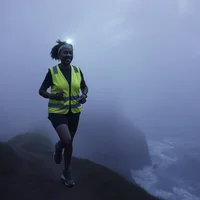 Woman in hi-vis vest and headlamp running along a foggy coastal cliff at dawn