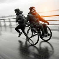 Person pushing a smiling wheelchair user at speed along a waterfront promenade, motion blur in background