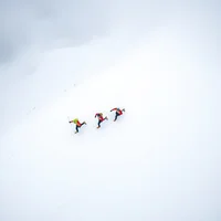 Three runners in colorful gear ascending a steep snowy mountain slope, viewed from above