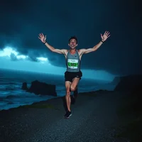 Runner raising both arms triumphantly on a coastal road under a dark stormy sky at dusk