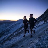Two adult para-athletes in monochrome gear run mountain switchbacks at twilight, captured as glowing light trails against a stark winter ridge.