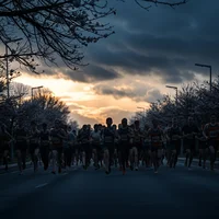Silhouetted barefoot race pack in reflective gear and headlamps on a blossom avenue beneath dramatic pre-dawn storm clouds.
