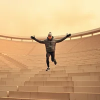 Elite runner in winter layers bounding up vast stadium steps through amber wildfire smoke haze on a summer morning.