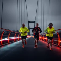Three runners in neon yellow and black jackets jogging across a suspension bridge with red lit cables in overcast weather.