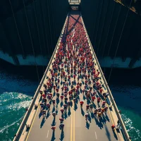 Overhead view of marathon runners crossing suspension bridge, colorful crowd forming flowing patterns in harsh midday sun
