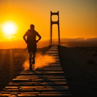 Silhouetted runner heading toward a suspension bridge along a path at sunset with orange sky