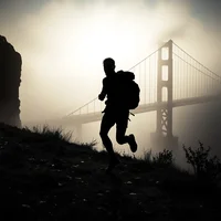 Silhouette of a backpack-wearing runner on a hillside with a large suspension bridge shrouded in fog behind them