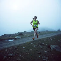Runner in a neon yellow tank top and dark shorts running on a misty gravel trail with rocks and fog behind