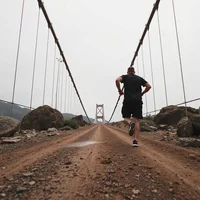 Runner in dark top and shorts seen from behind on a dirt road leading toward a suspension bridge under overcast sky