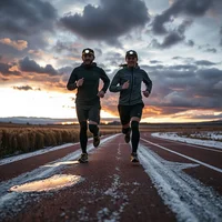 Two runners in dark jackets and caps running toward the camera on a red track with snow at sunset.