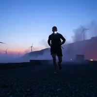 Silhouetted runner jogging on gravel at dusk, with wind turbines and a glowing building against a purple sky.