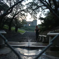 A person standing beside a water fountain on wet stone steps, with large trees and a stone tower at dusk