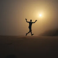 Silhouette of a person jumping with arms raised against a hazy golden sunset over flat sandy ground