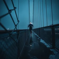Runner on a wet suspension bridge walkway at dusk, surrounded by metal cable fencing under a dark blue sky