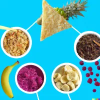 Overhead view of bowls of granola, pink fruit purée and banana slices, with berries, a banana and a pineapple on blue