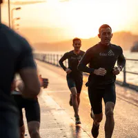 Three runners in dark athletic wear jog along a waterfront promenade beside a railing at golden sunset.