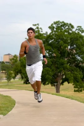 Man in grey tank top and white shorts running along a paved path past green trees and grass