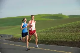 Two runners jogging side by side on a rural road past rolling green hills under a pale sky.