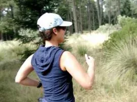 Person in a light cap and navy sleeveless hooded top outdoors on a grassy trail among pine trees