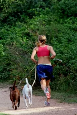 Woman in red sports bra and blue shorts jogging on a dirt path with two dogs on leashes, trees behind her.